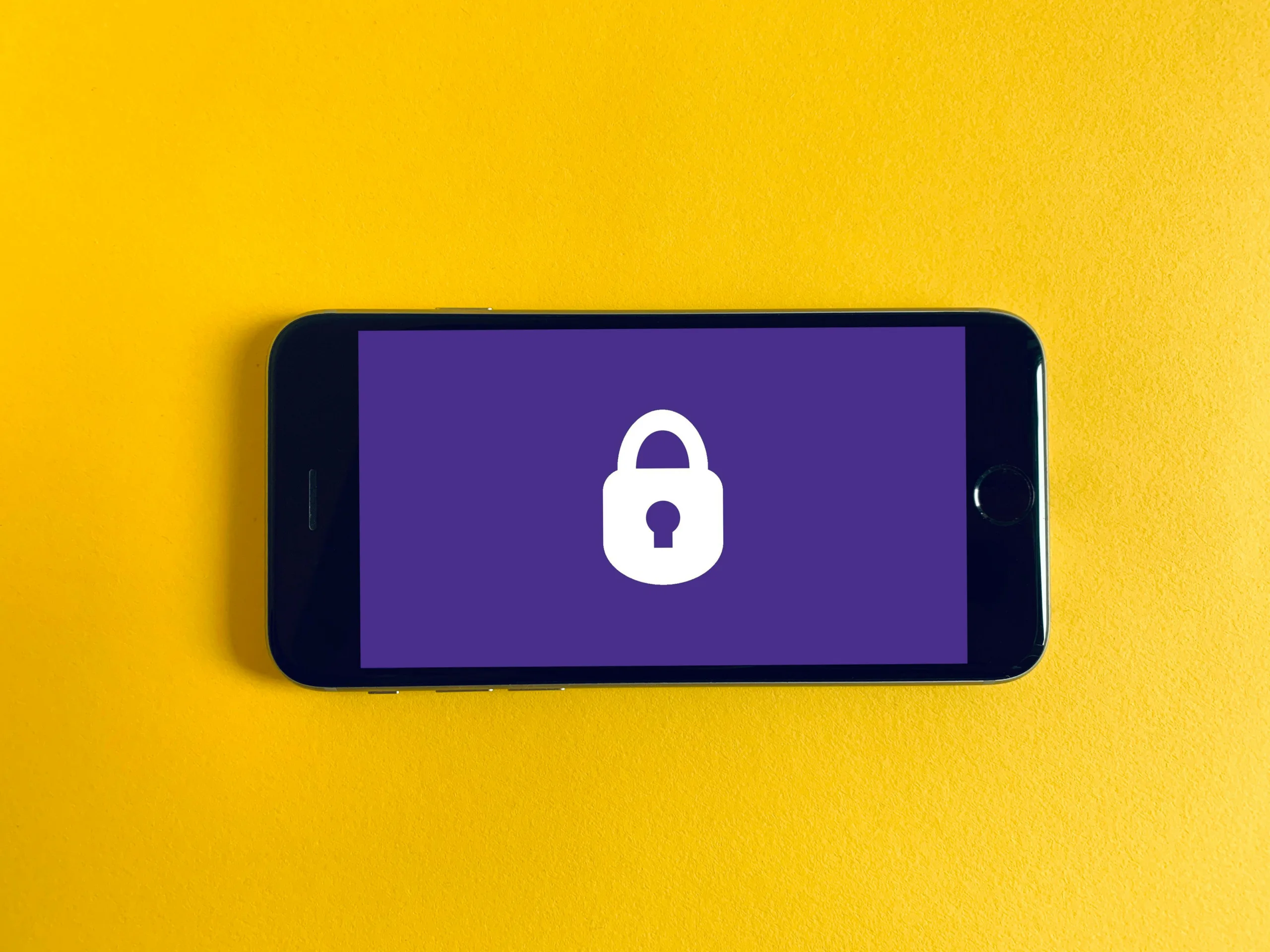 Smartphone with a screen displaying a white padlock icon on a purple background, placed on a bright yellow surface