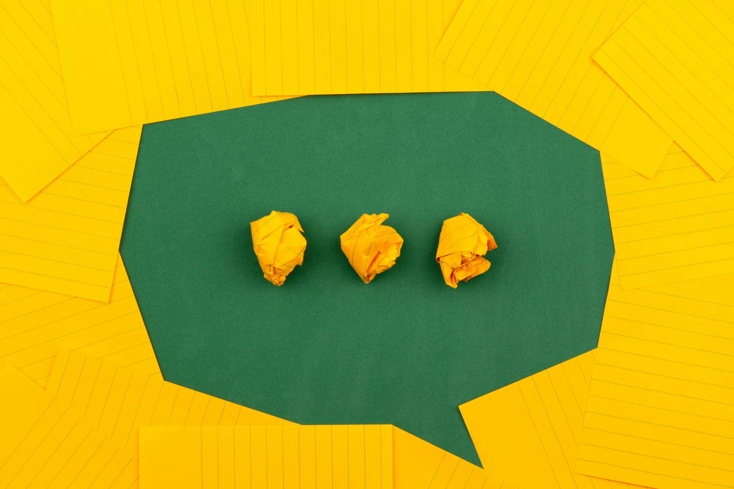 Yellow lined papers arranged around a green speech bubble shape, with three crumpled yellow paper balls inside the speech bubble.