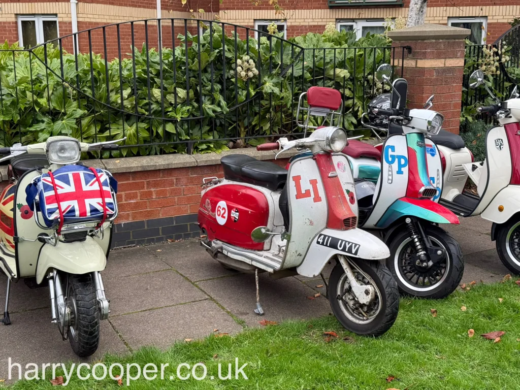 Close-up of a row of vintage scooters, each decorated uniquely, including one with a Union Flag bag on the front and another with "LI" in red on a white background. The scooters are parked near a brick wall and foliage.