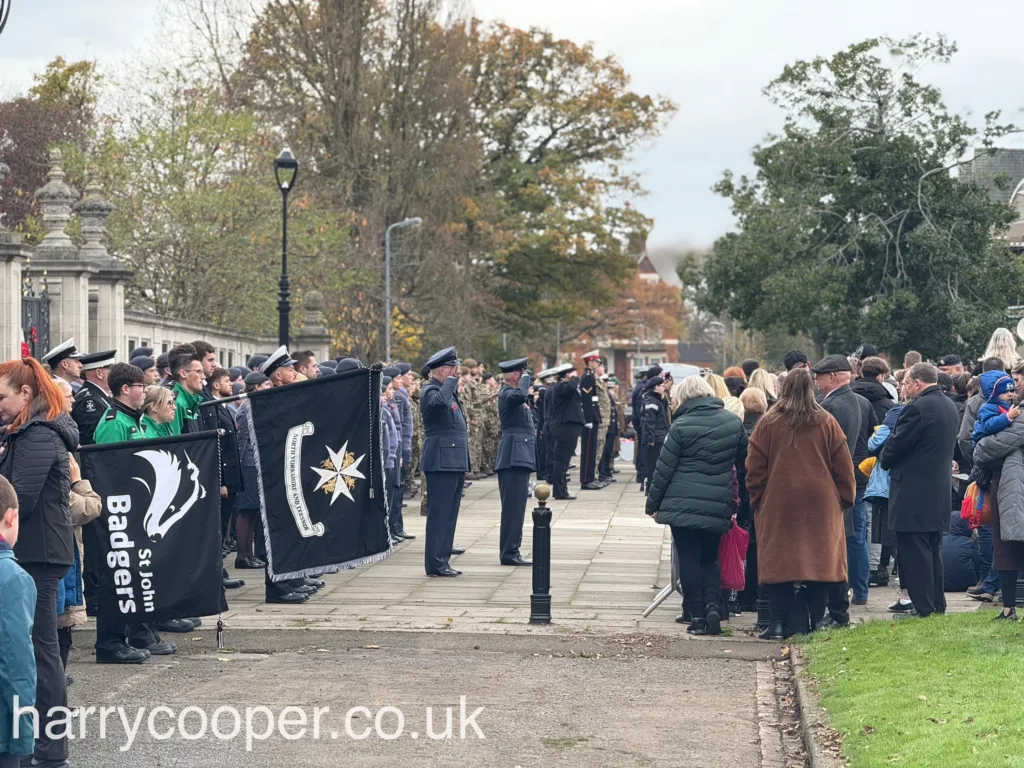 People gathered by a park walkway, with uniformed cadets holding flags, including one with the "St. John Badgers" logo. Other attendees stand nearby, observing the ceremony.