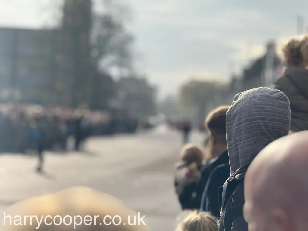 A blurred shot of a crowd of spectators standing along the roadside with the back of 1 persons head in focus, creating a hazy atmosphere with the sunlight filtering through, emphasising the depth of the crowd waiting for the procession.