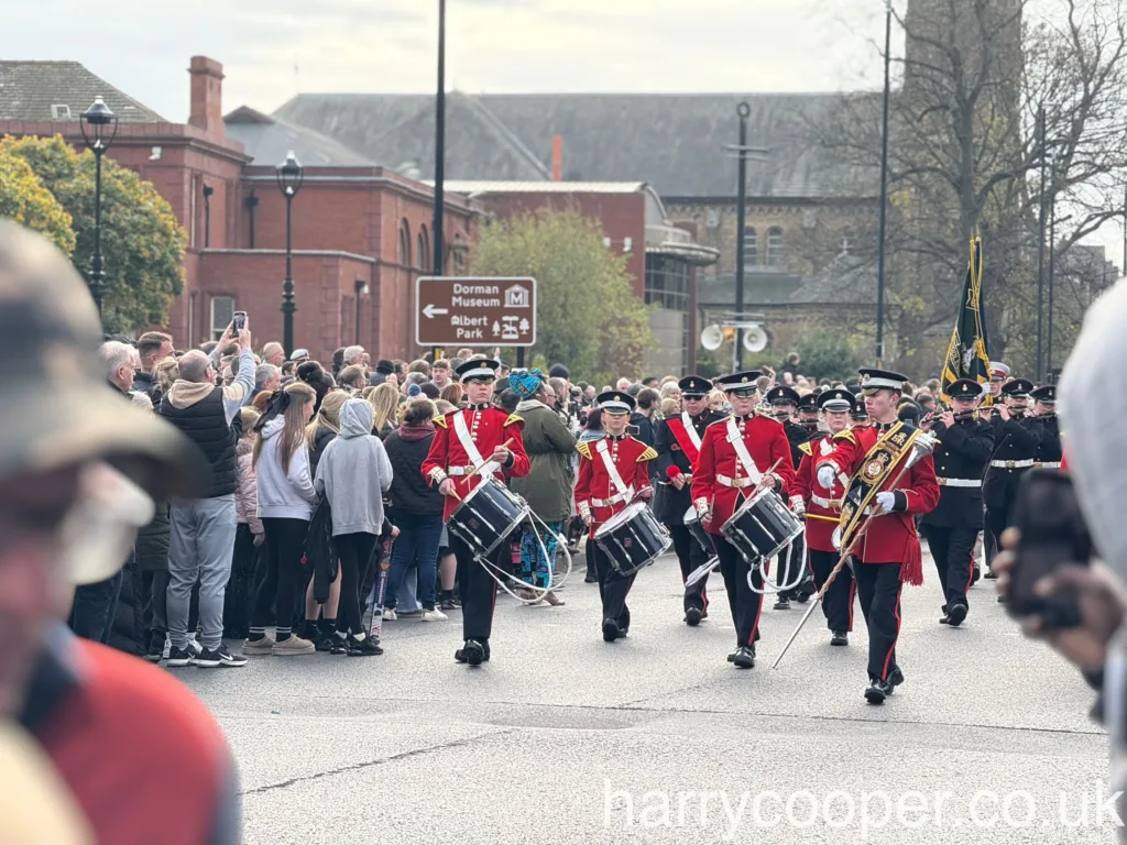 A wide view of the marching band in red and black uniforms, playing their drums and marching down a street lined with spectators, including children and families observing the procession.