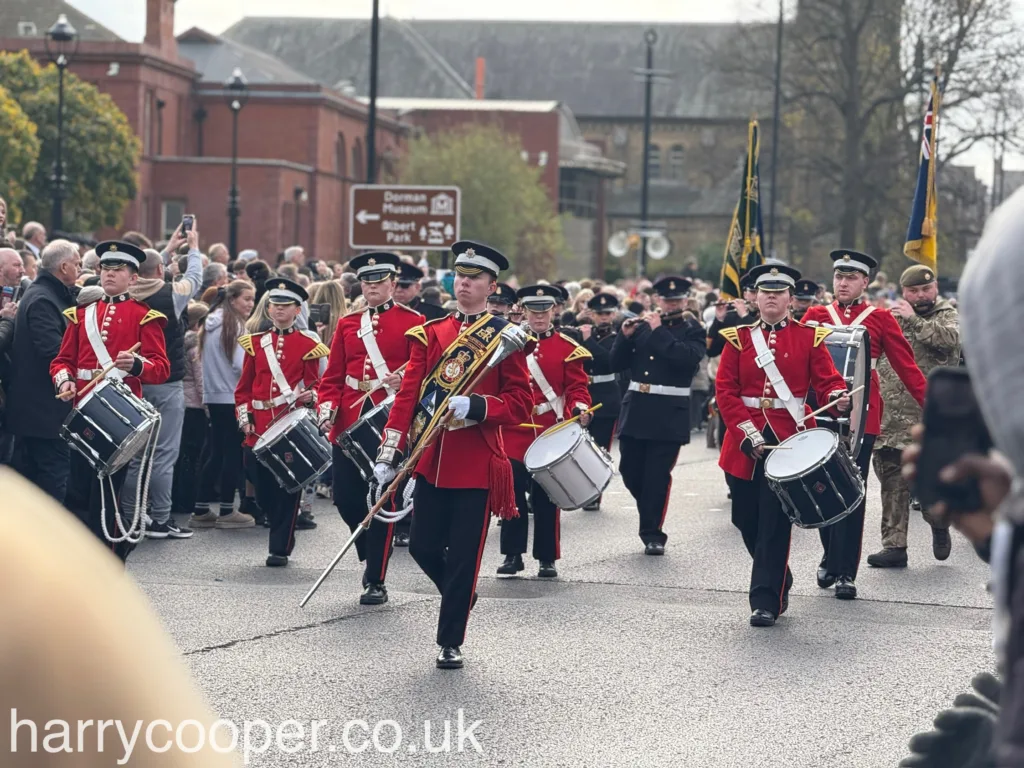 A marching band dressed in red and black ceremonial uniforms, led by a young man holding a large ornate baton. The band members carry drums, and the crowd lines the street watching their performance.