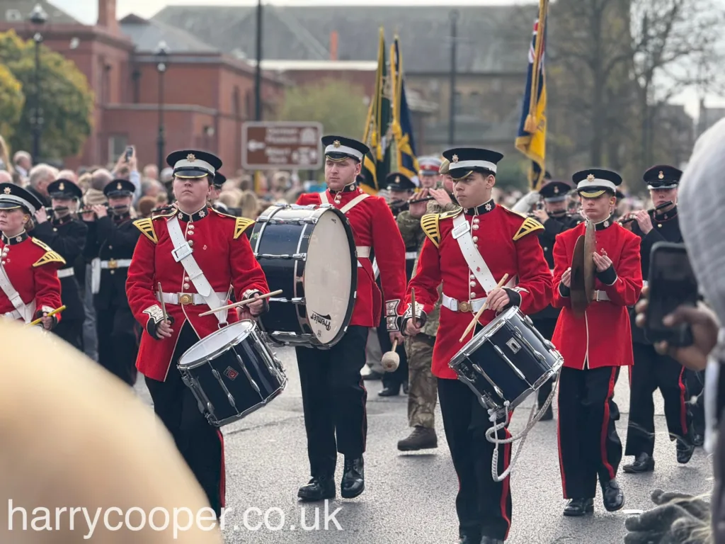 A group of young drummers in red and black ceremonial uniforms marching down the street. They carry large drums and wear black hats, accompanied by a crowd of spectators on both sides of the street.