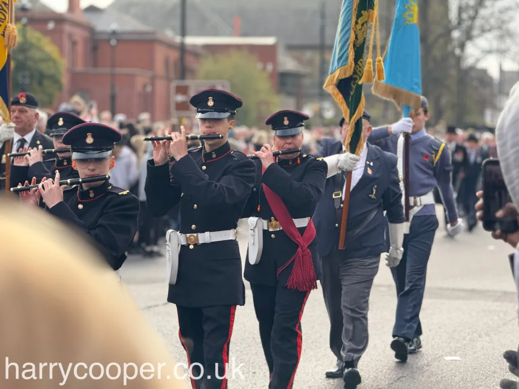 A band of military personnel in black uniforms play flutes as they march. They are led by a banner bearer, and the crowd observes the performance as part of the Remembrance Day event.