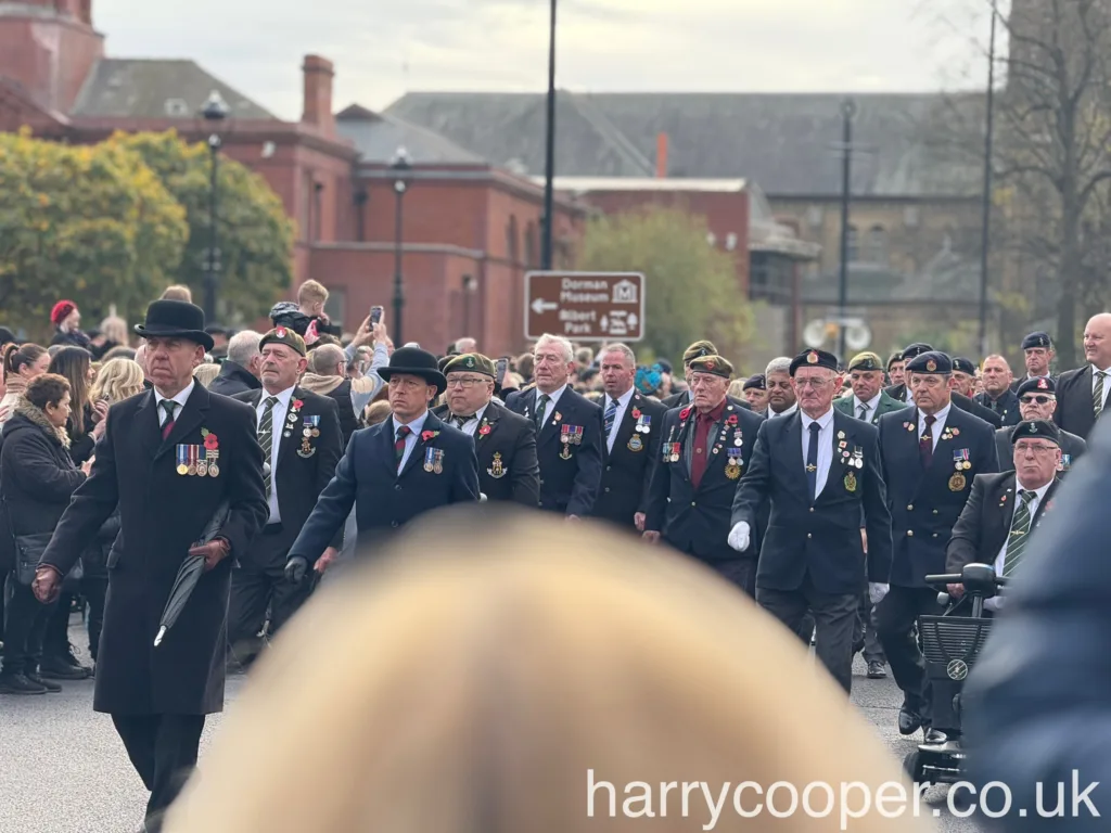 Veterans in black blazers, some wearing traditional bowler hats, walk in the parade, displaying medals on their jackets as they participate in the Remembrance Day procession.