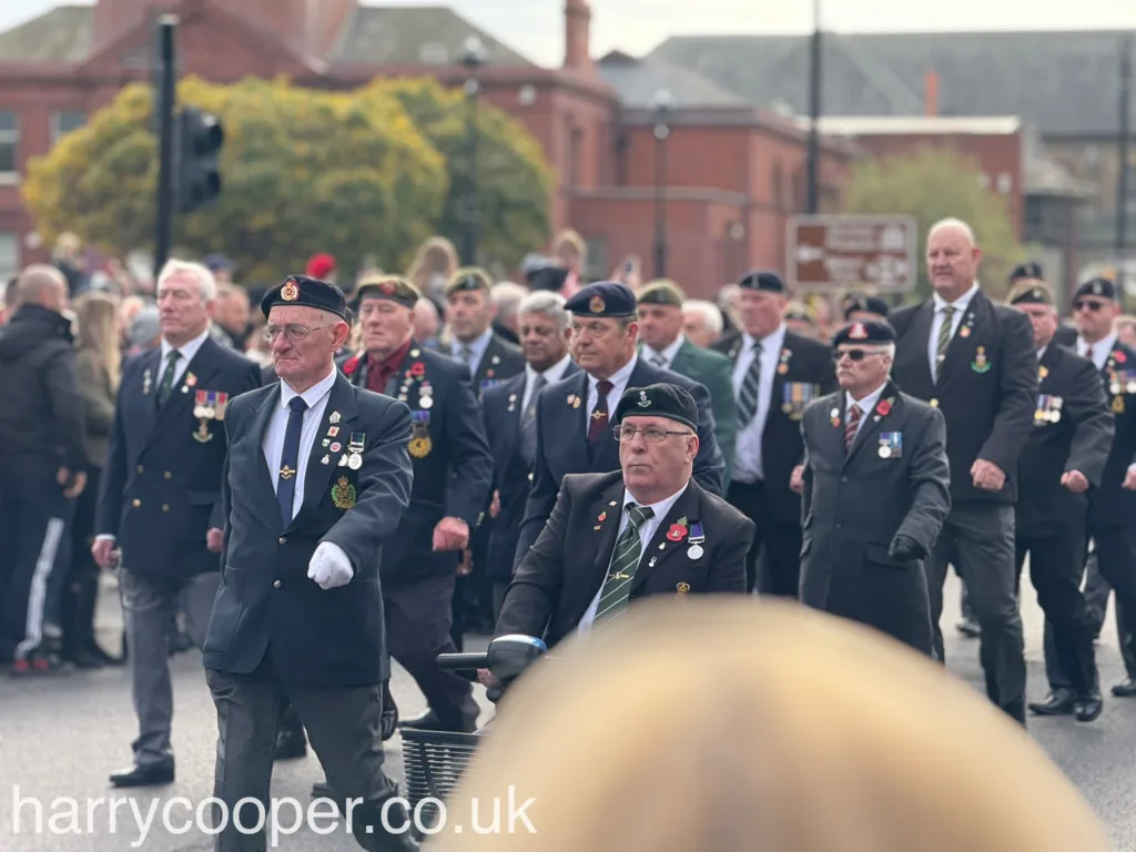 Veterans dressed in black blazers, some with berets, march alongside one another, wearing medals and poppies. The crowd watches and honours their dedication on Remembrance Day.