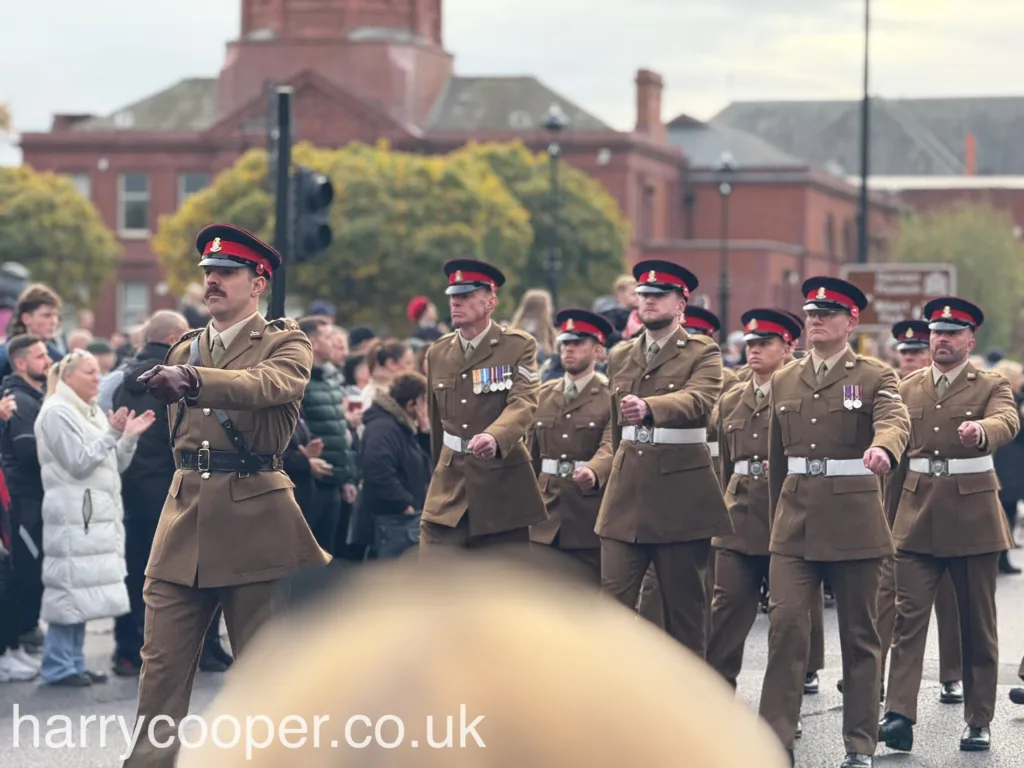 A group of military personnel in tan dress uniforms, marching with medals and poppies on their chests, observe Remembrance Day with reverence as they proceed through the crowd.