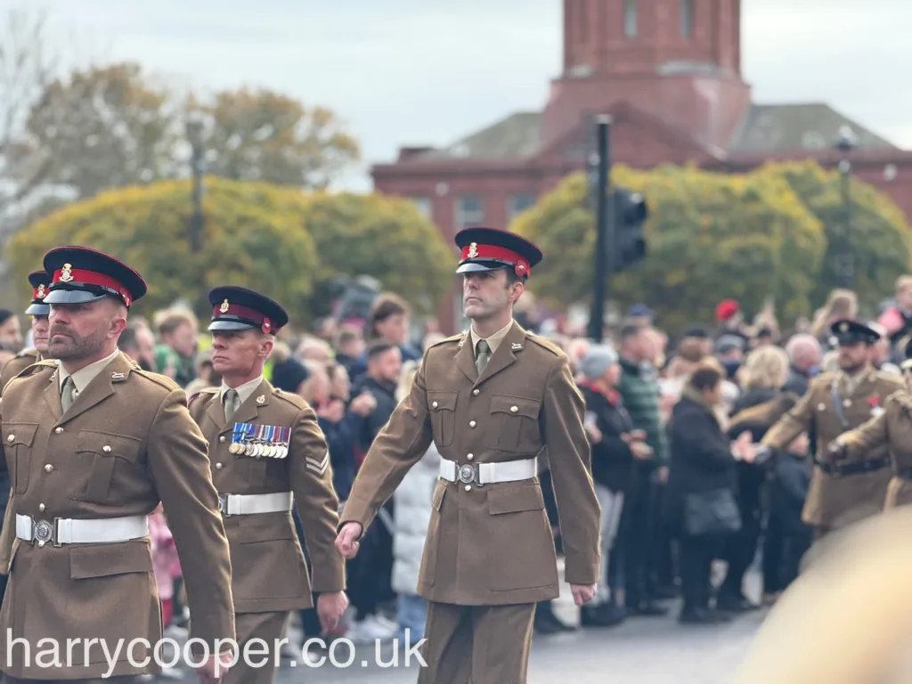 Military personnel in tan dress uniforms with red and black caps march in formation. Their attire includes white belts, and they display medals on their chests as part of the Remembrance Day service.