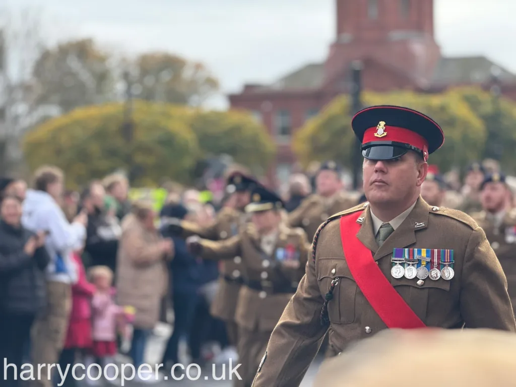 A close-up of a military officer in tan dress uniform with a red sash and an array of medals, marching with a solemn expression during the Remembrance Day event.