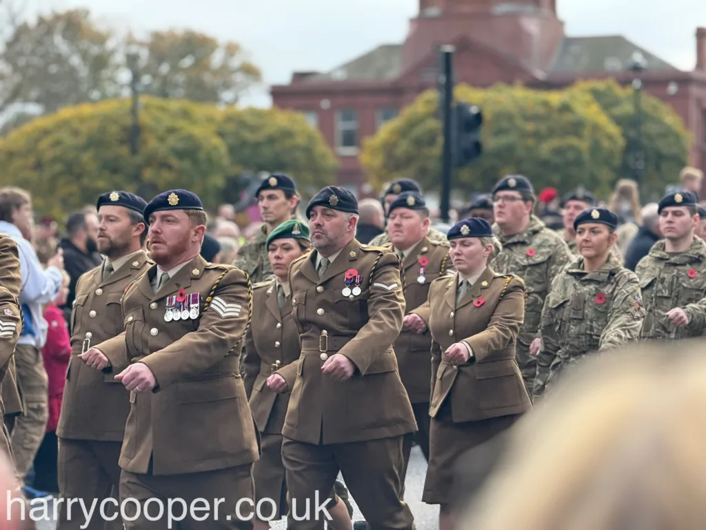 Soldiers in tan military dress uniforms adorned with medals and poppies march in unison, solemnly observing Remembrance Day. Their expressions reflect respect and remembrance.