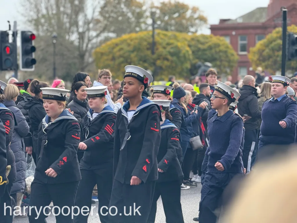 Sea cadets in traditional naval attire, including black uniforms with sailor collars and white hats, march in formation. They display poppies on their uniforms as part of the Remembrance Day procession.