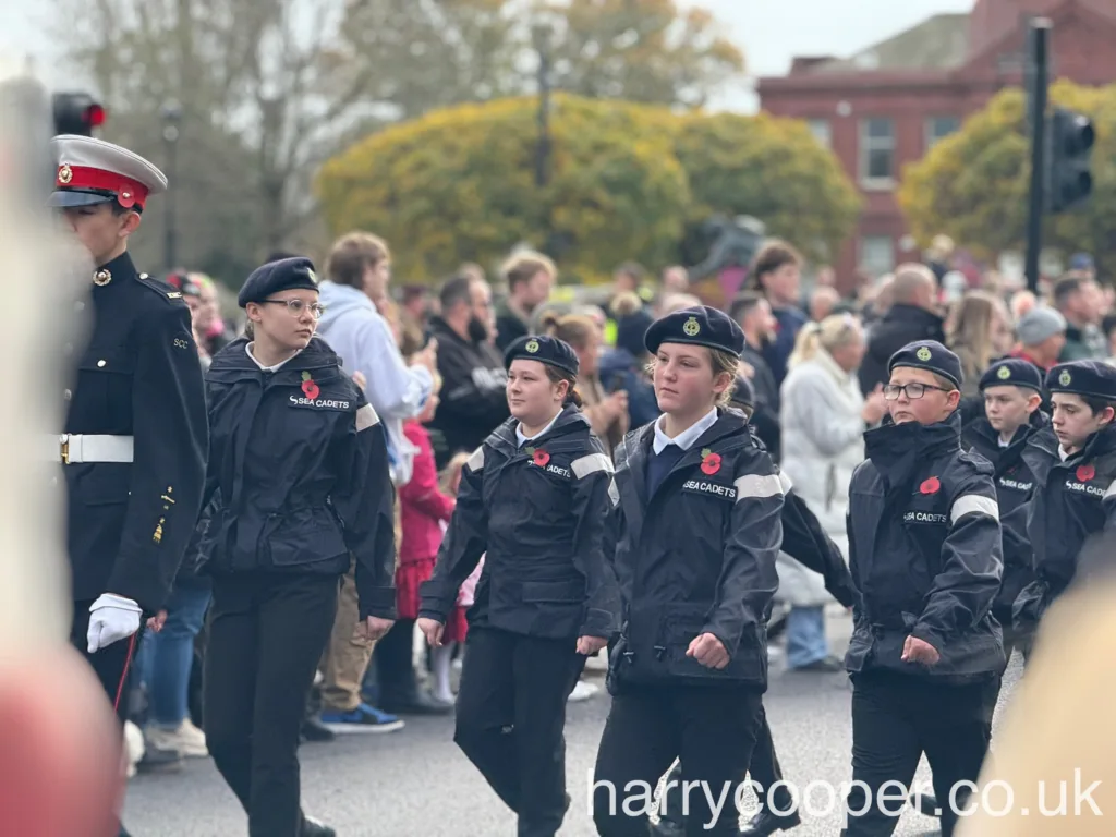 A group of young sea cadets in black uniforms and berets march in formation, with one cadet in a red and white military hat leading the group. They wear poppies on their uniforms in honour of Remembrance Day.