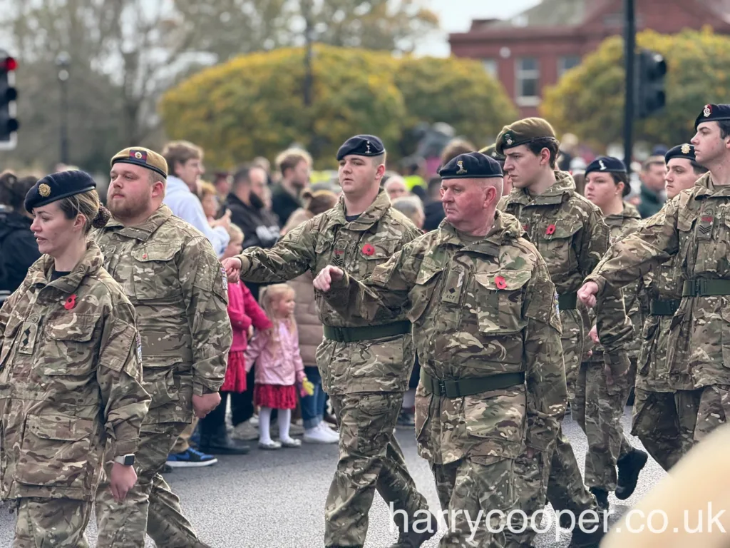 Cadets and adult officers in camouflage uniforms, marching in unison with poppies, commemorating Remembrance Day, with onlookers in the background.