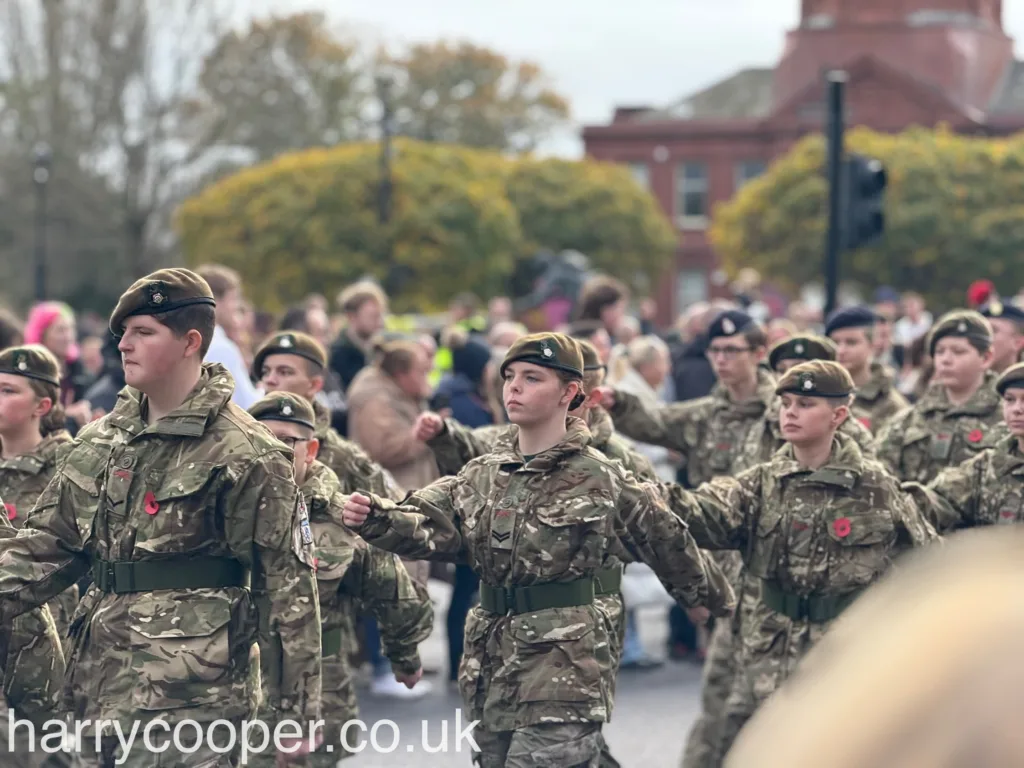 A group of young cadets in camouflage uniforms and berets march in formation during the Remembrance Day parade. Each cadet wears a red poppy on their uniform in commemoration, and they march with their arms outstretched in synchronisation.