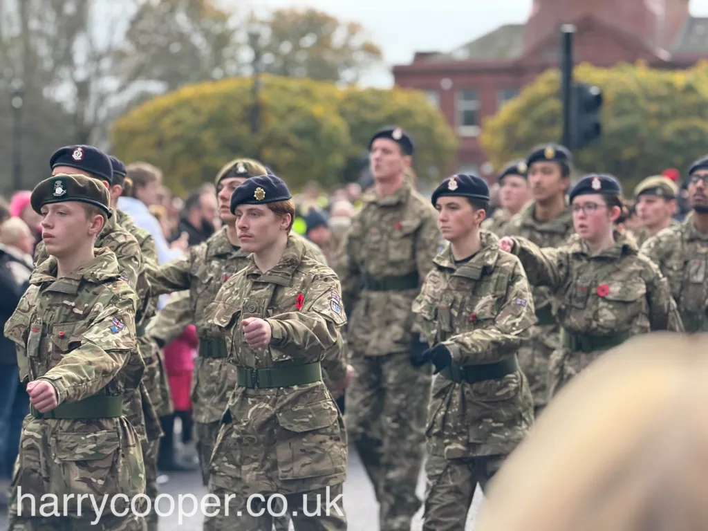 A close-knit formation of cadets in camouflage uniforms, marching in line with poppies on their jackets, against a backdrop of watching spectators.