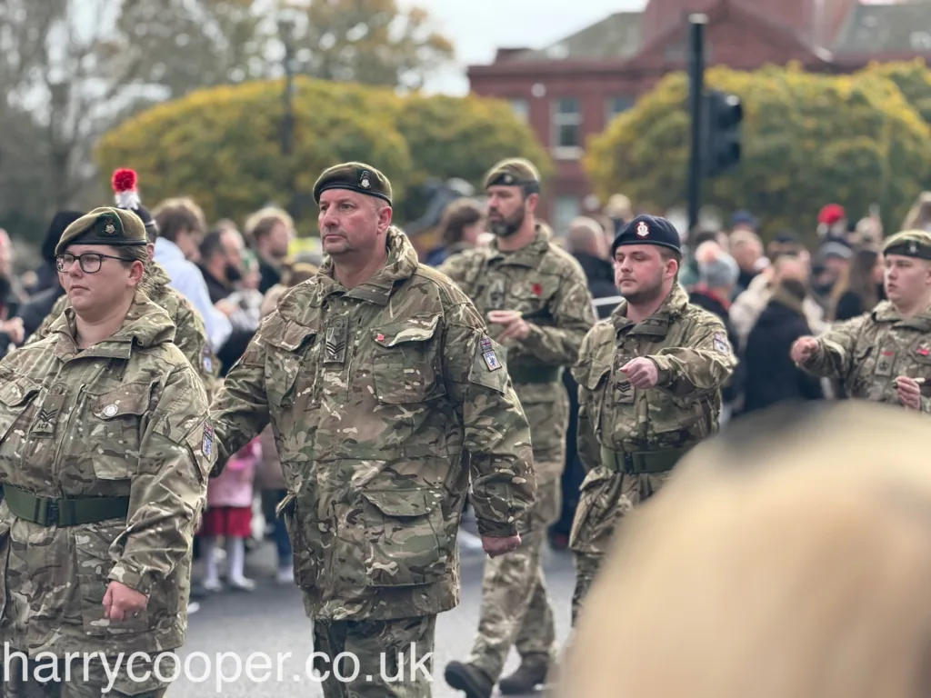 A group of adult and young cadets in camouflage attire, marching in formation during the Remembrance Day parade, with children in the background.