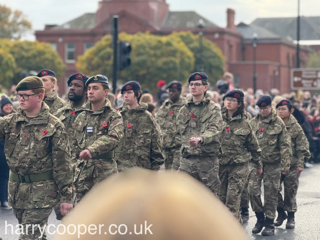 A formation of army cadets in camouflage uniforms, marching in unison with red poppies on their jackets, honouring Remembrance Day.