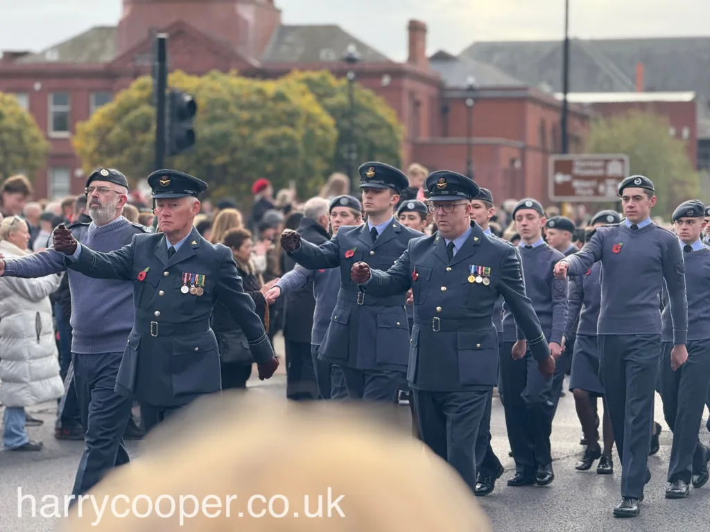 A group of senior officers in blue uniforms marching with arms raised in a formal march, adorned with medals and poppies, with cadets marching behind them.