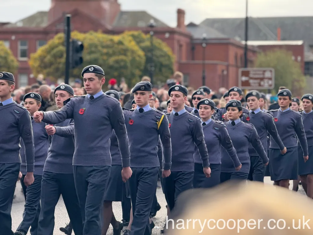 Rows of cadets in blue uniforms with berets and red poppies, marching in unison with focused expressions, with spectators lining the street.