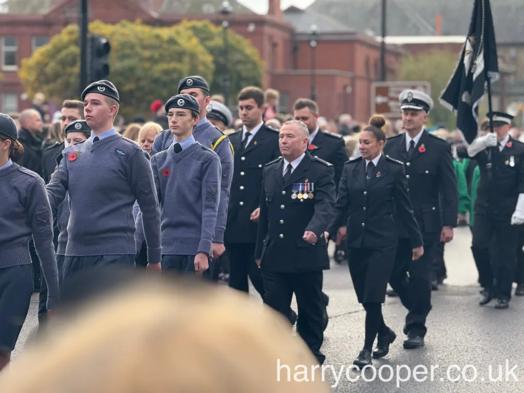 A group of cadets in blue uniforms marching in formation, wearing berets and red poppies on their chests, with a crowd of spectators in the background.