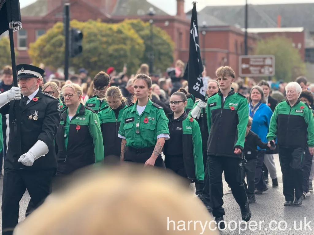 A group of St. John Ambulance members, wearing green and black uniforms, march in formation during the remembrance day parade, led by a senior officer holding a flag.