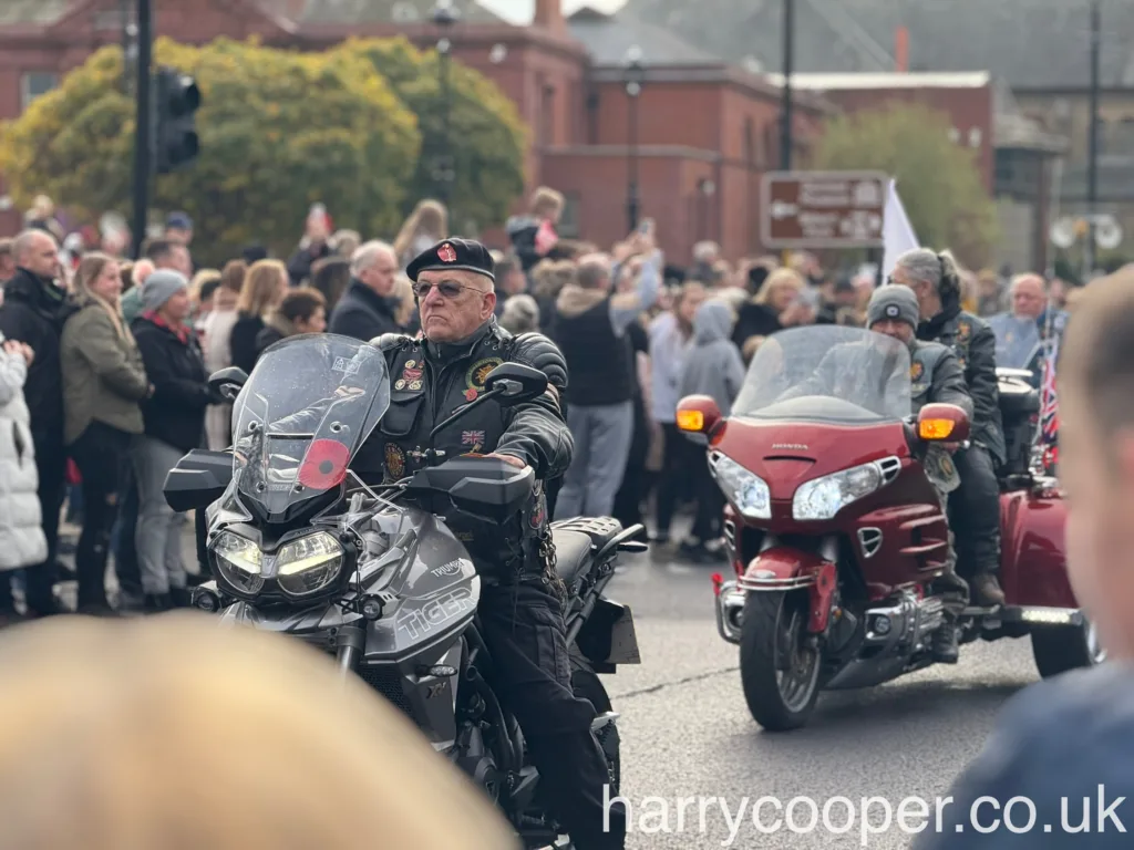 A veteran, wearing a black beret and covered in military patches, rides a grey motorcycle with a poppy decal in a remembrance day parade.