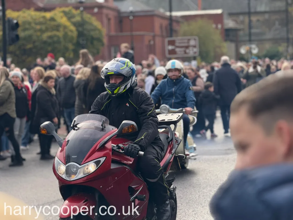 A motorcyclist on a red motorcycle with a black helmet and jacket, with a dense crowd in the background.