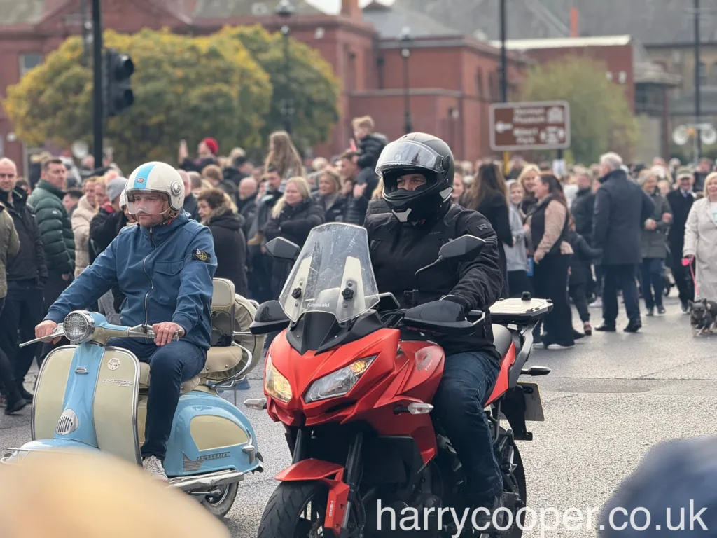 A motorcyclists on a red motorcycle, and a scooterist on a light blue and white scooter, ride side-by-side during a remembrance parade. The crowd watches from behind.