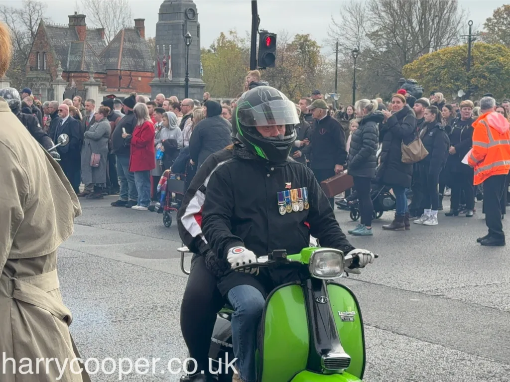 A scooterist with a green scooter decorated with medals rides in the procession, watched by attendees.