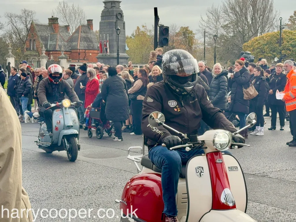 Several scooterists in a parade, riding scooters, with a large crowd of onlookers.