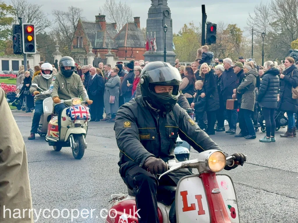 A close-up of a scooterist in a black jacket and helmet on a red and white scooter, adorned with an "LI" sticker and a poppy sticker, as part of a remembrance procession with a crowd in the background.