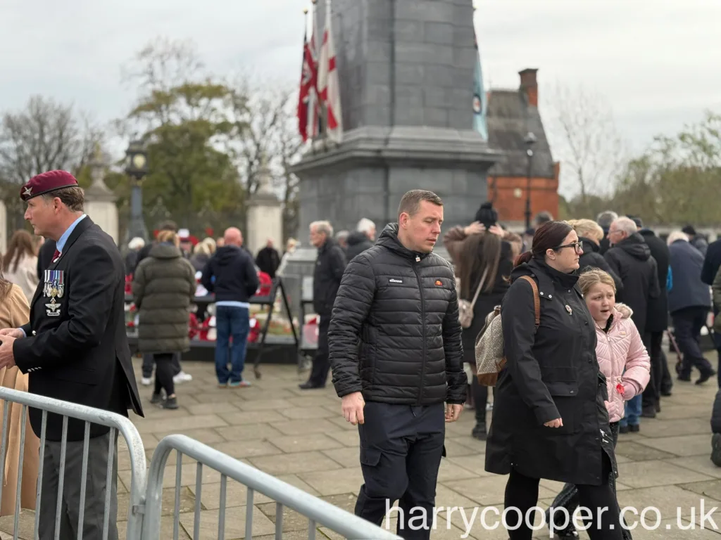 Attendees in winter coats stand around the cenotaph, some placing poppies on memorial stands.