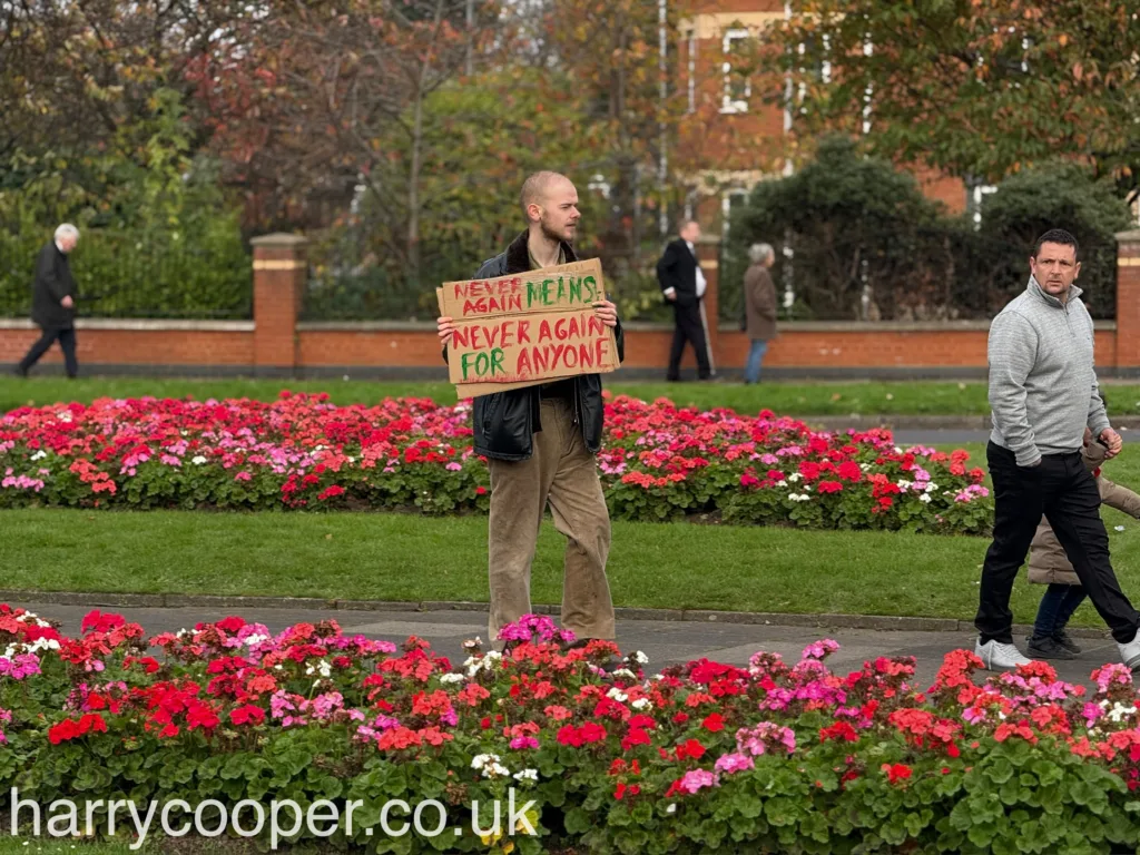 A man stands holding a handmade sign reading “Never Again Means Never Again for Anyone,” advocating for peace and remembrance, pointing at the camera.