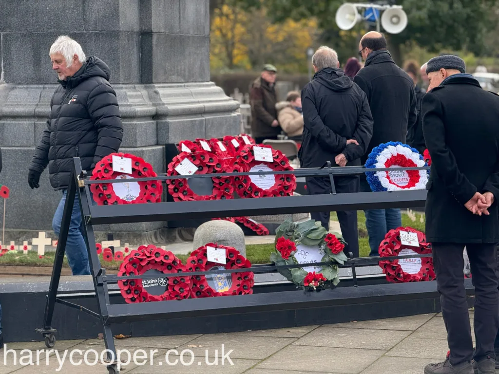 Red and blue poppy wreaths from various organisations are displayed on metal stands beside the cenotaph as people pass by.