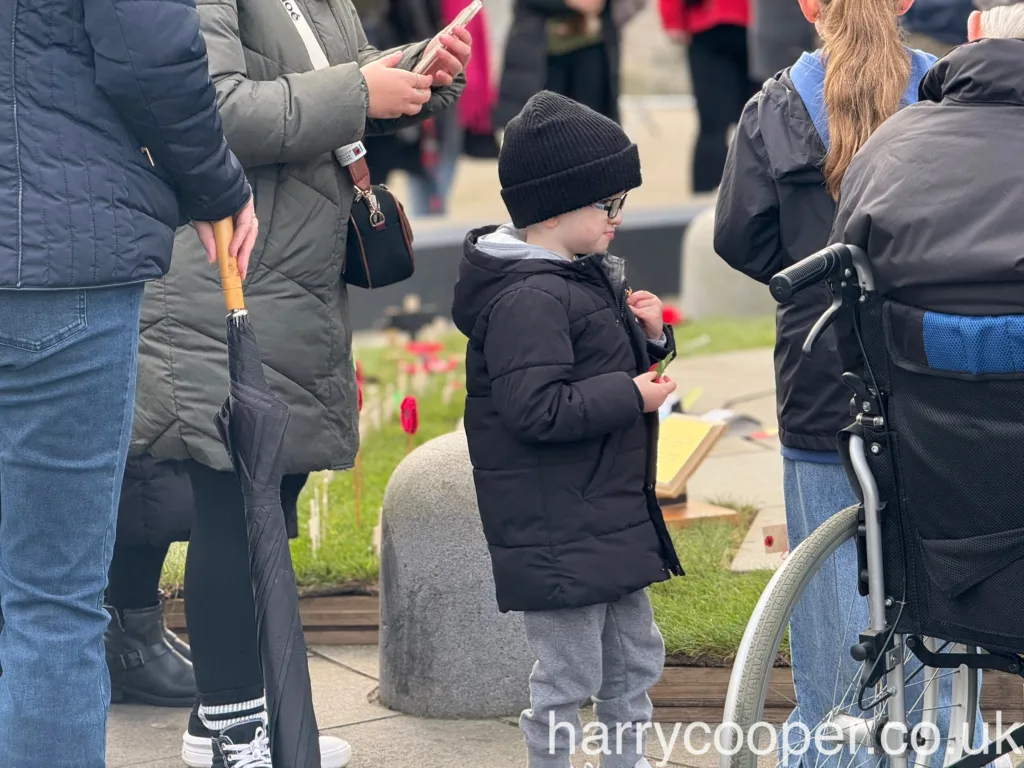 A young child, bundled in a black jacket and hat, holds a single red poppy while standing near the memorial.