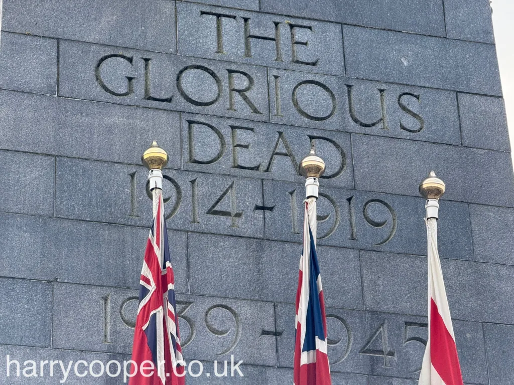 Close-up of British flags flying beside a stone inscription reading “The Glorious Dead 1914-1919, 1939-1945” on the cenotaph.