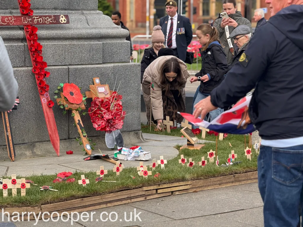 A group of people, including a woman placing a small cross, pays their respects beside poppy-adorned wooden crosses and red floral tributes at the cenotaph.