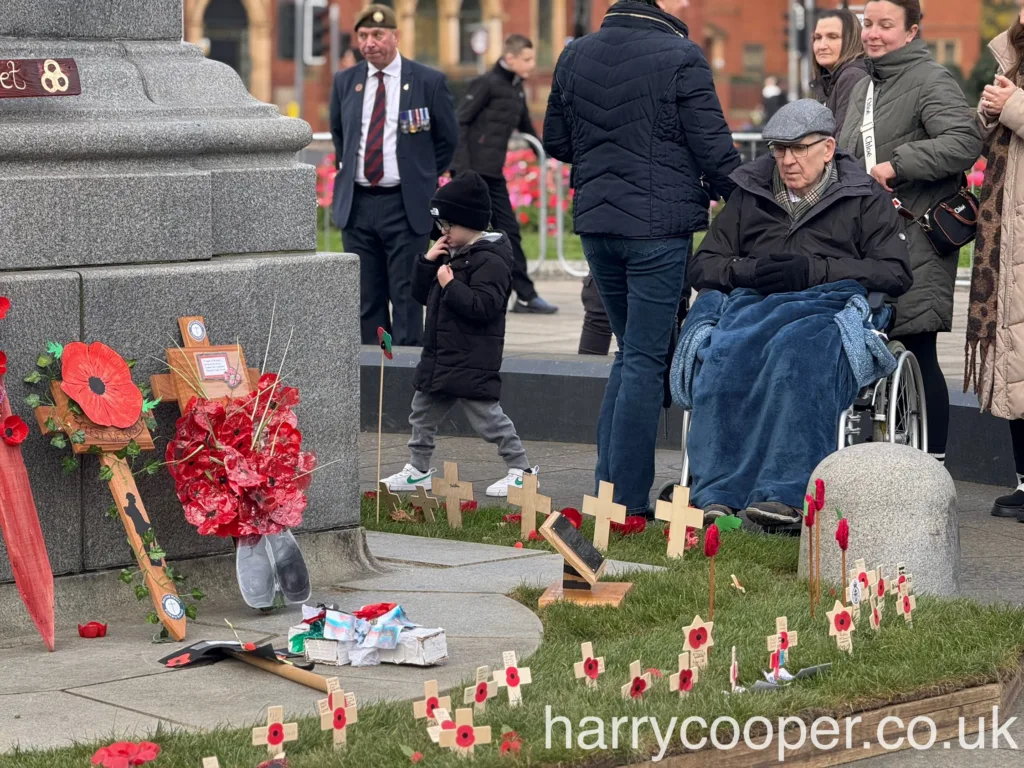 An elderly man wrapped in a blue blanket sits in a wheelchair near small wooden poppy crosses on the grass, with a young child and others nearby in quiet reflection.