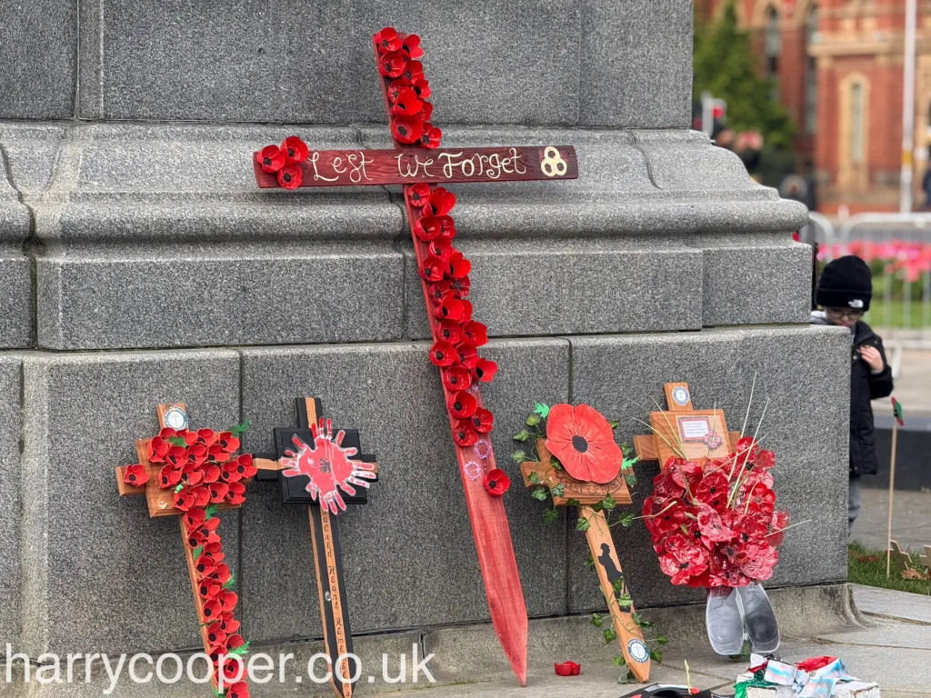 A display of wooden crosses adorned with red poppies stands against the grey stone of the cenotaph, with the words "Lest We Forget" written on a large red cross.