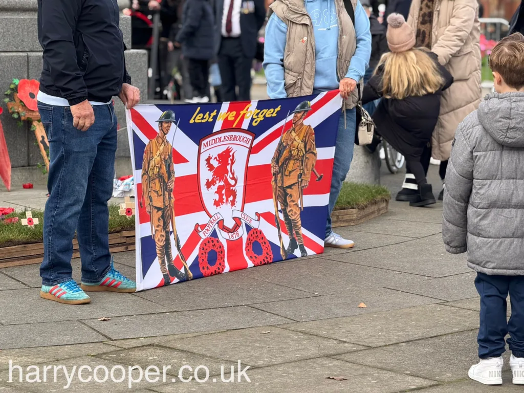 Two people hold a banner with the Middlesbrough crest, poppies, and images of soldiers, with the words "Lest We Forget" at the top.