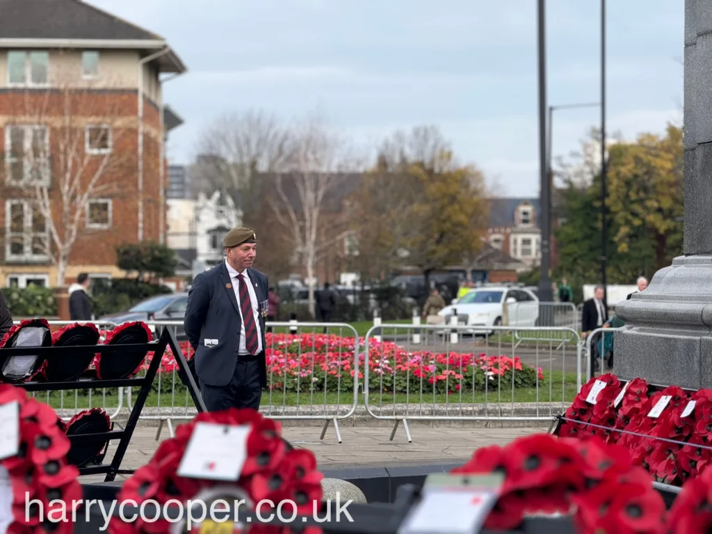 A veteran wearing a military beret and medals stands beside rows of red poppy wreaths, with a flower bed and the cenotaph in the background.