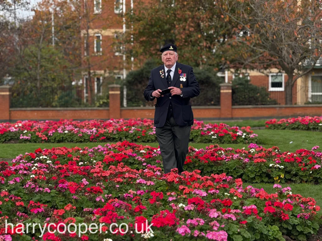 An older veteran, wearing medals on his blazer, stands solemnly in a garden bed of red and pink flowers.