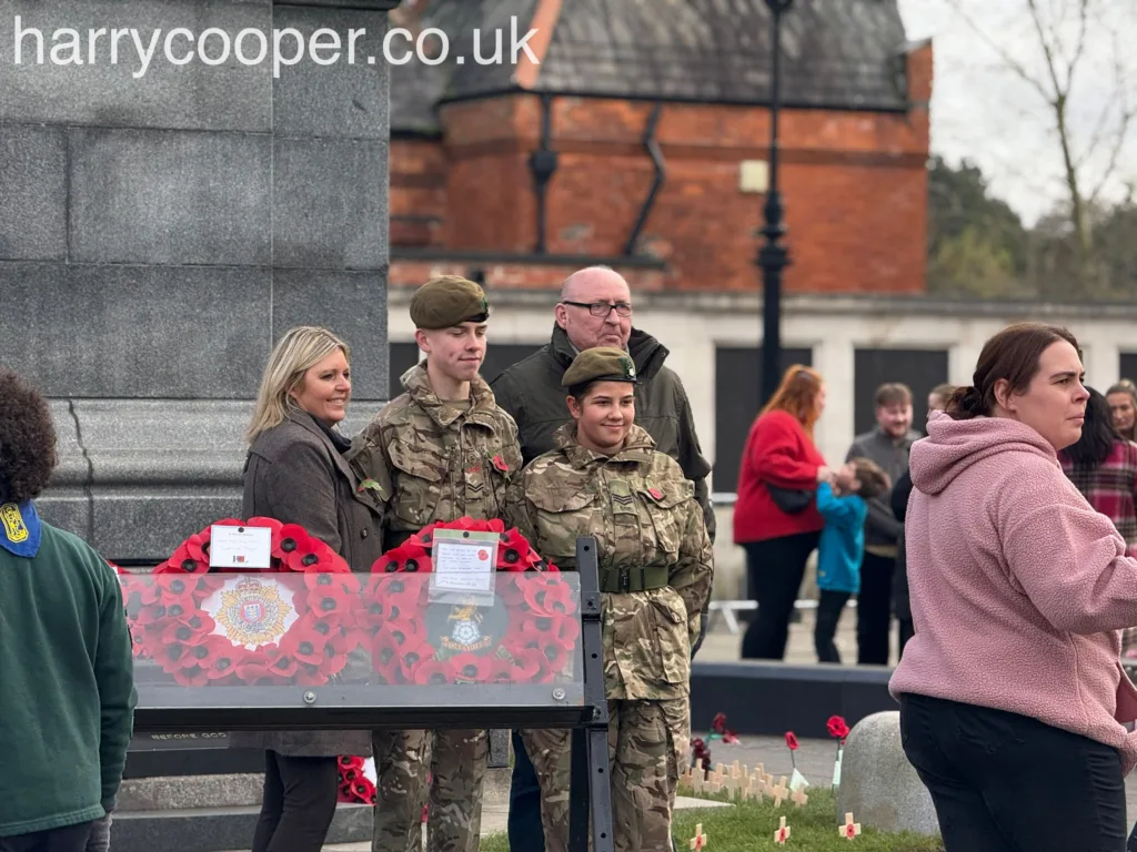 A group of people, including two young soldiers or cadets in uniform and two adults, stands near red poppy wreaths laid at the cenotaph.