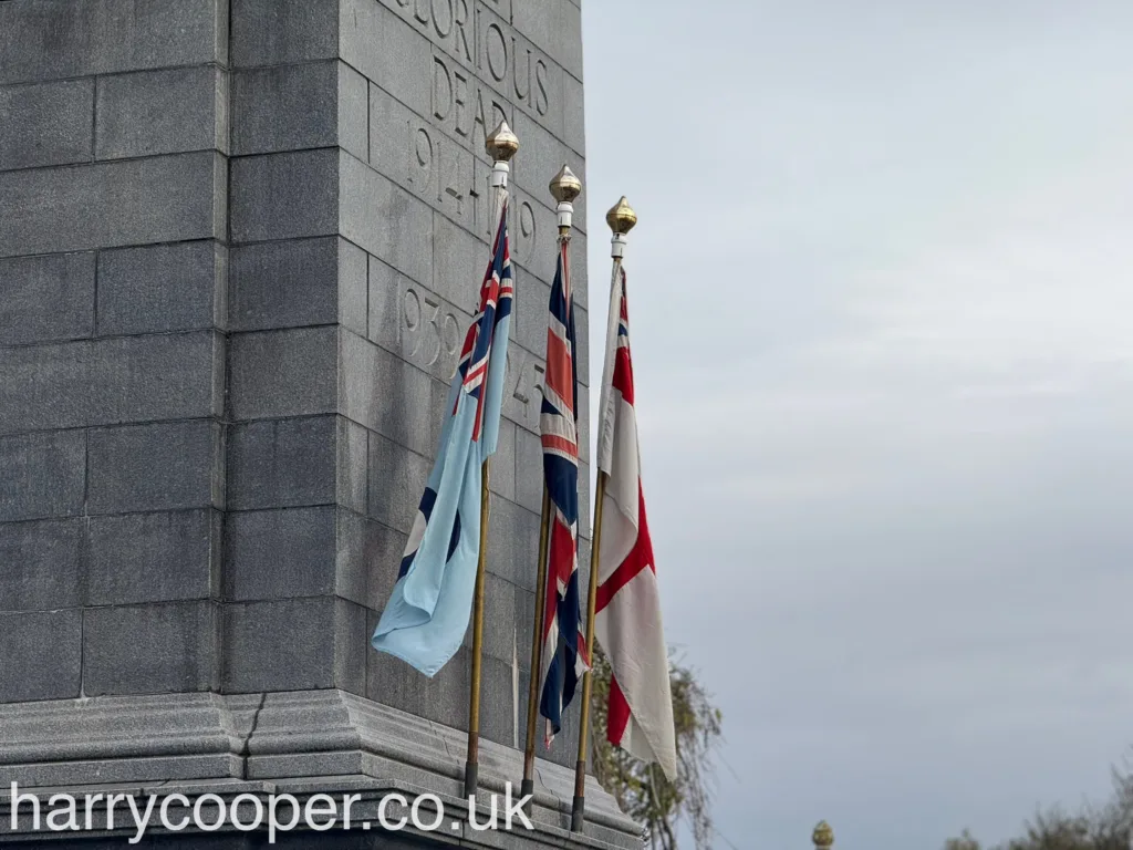 A close-up of flags, including the Union Flag, RAF flag, and the Saint George's Cross, flying beside a grey stone cenotaph engraved with the words “The Glorious Dead 1914-1919, 1939-1945.