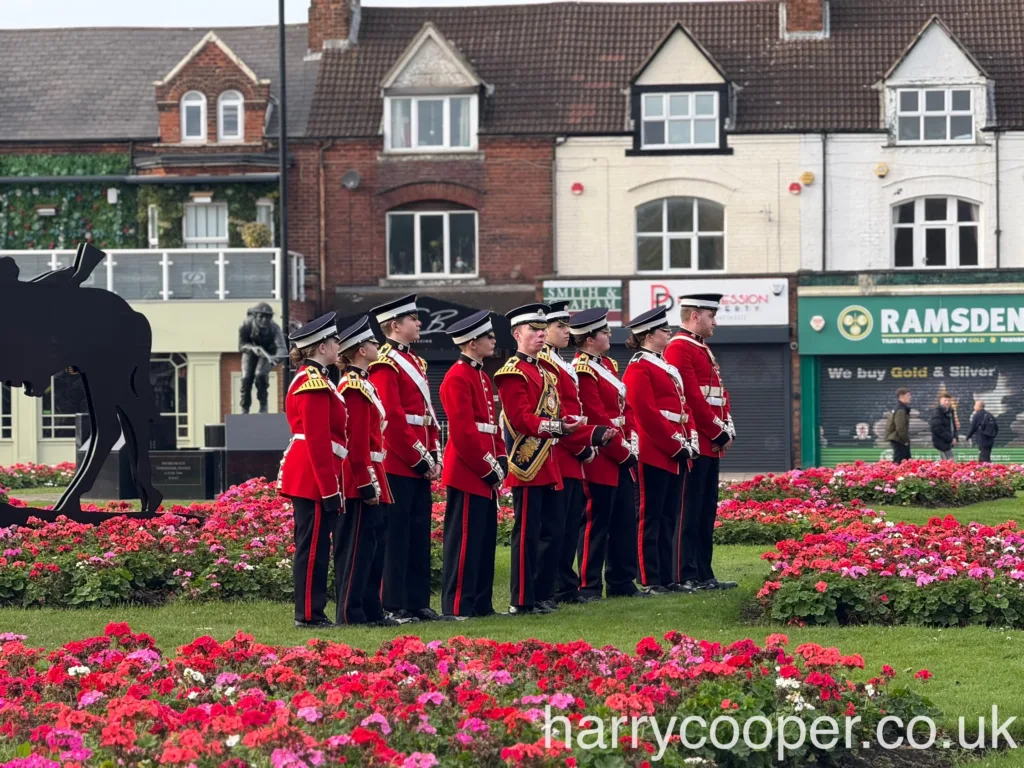 A line of soldiers in red uniforms stands at attention in a garden filled with vibrant flowers, with silhouettes of soldiers and civilians in the background.